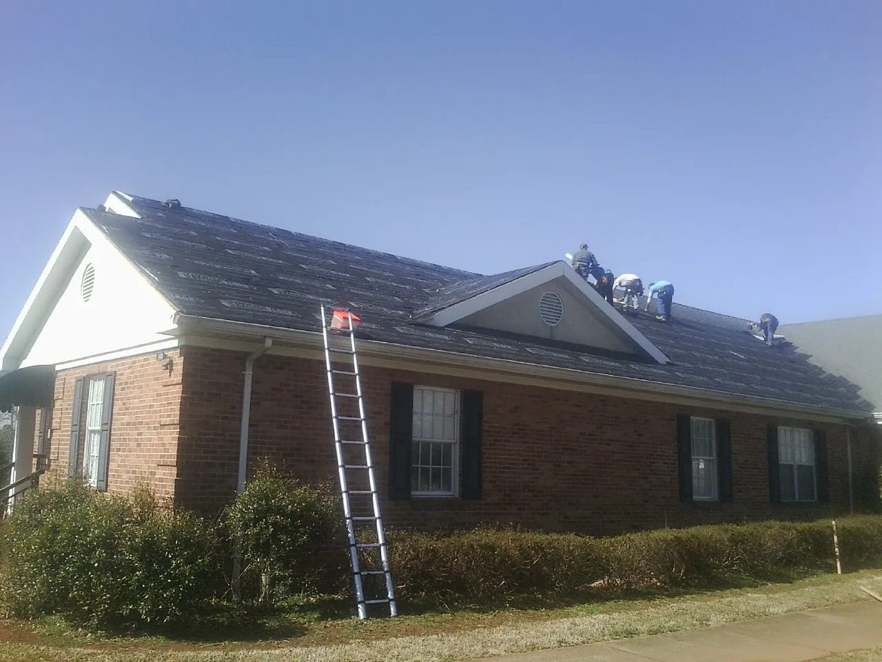 Crew tearing shingles off a brick ranch roof with a ladder set — Emergency Roof Repair work by our crew in Lorimor