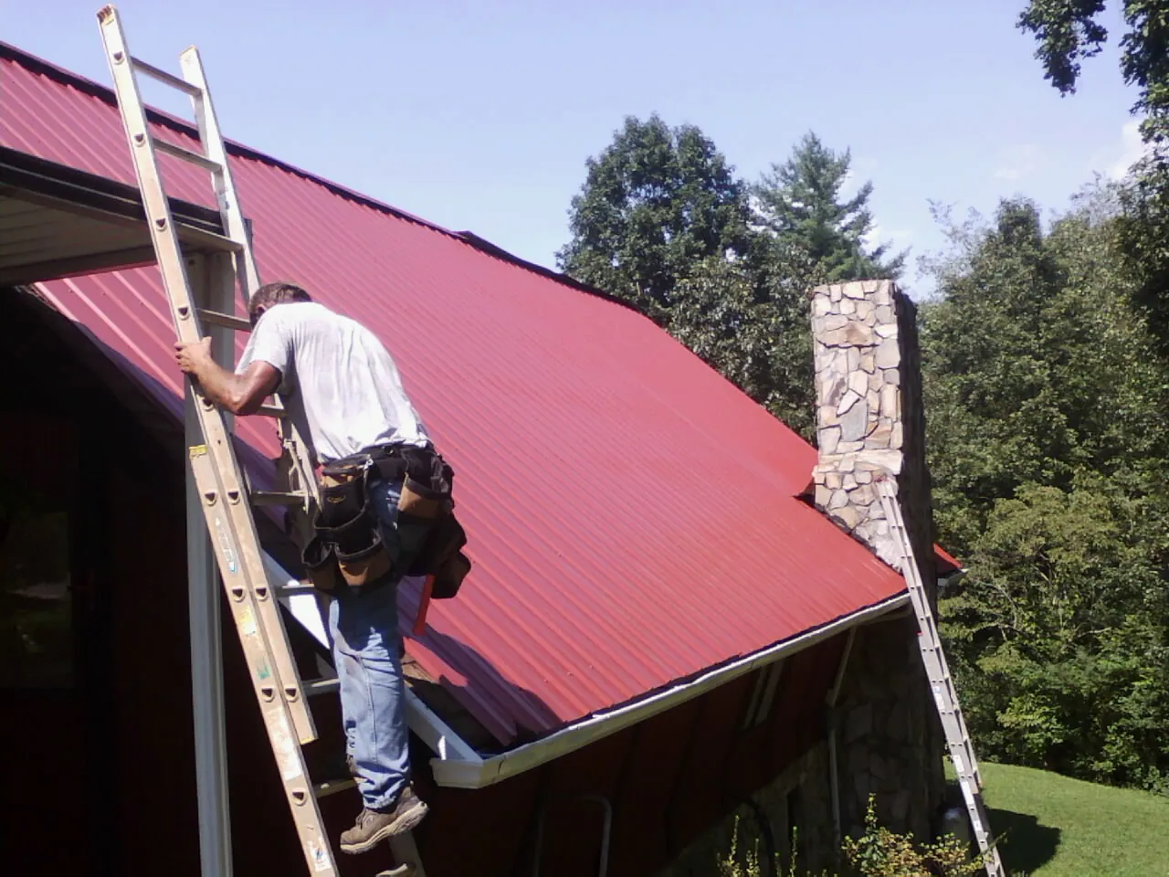 Roofer on a ladder beside a red metal roof and stone chimney — Soffit & Fascia Repair by Roofing Brothers in Lorimor